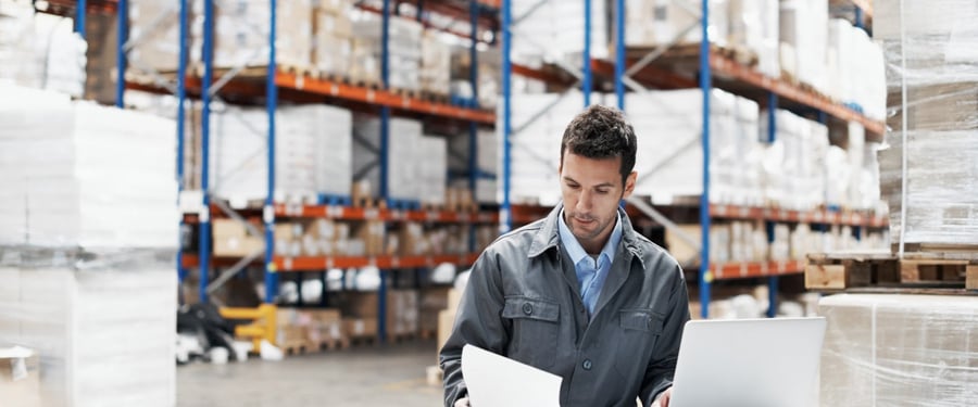 A young man using his laptop and looking over his notes while working in a warehouse